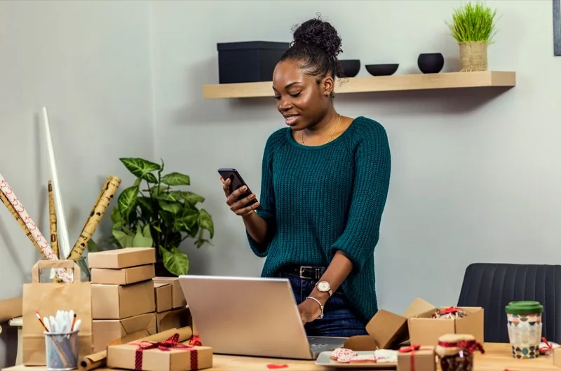 A founder checking her phone beside a laptop, surrounded by packed orders ready to ship