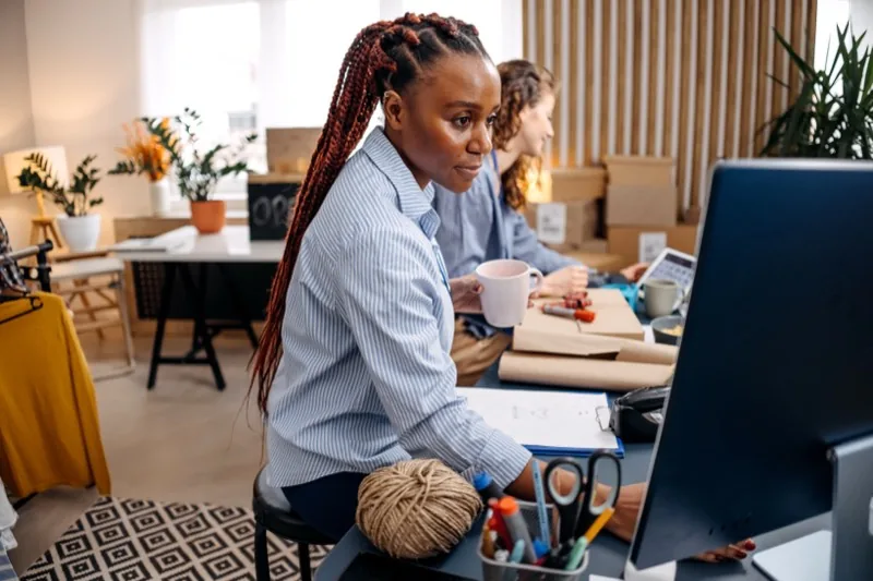 A founder at her desk in a shared studio workspace, a colleague working behind her